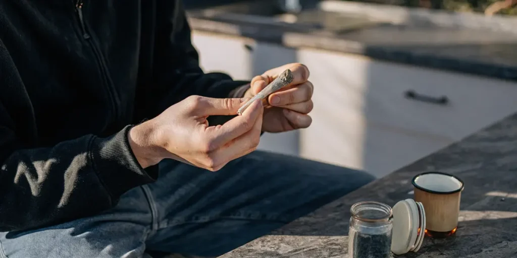 Hyper-realistic scene of a person's hands finishing rolling a cannabis joint, with a jar of cannabis and a mug on an outdoor table.