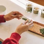 Hyper-realistic scene of a person's hands rolling a cannabis joint with ground material on a rolling paper, on a bright table with jars of cannabis.