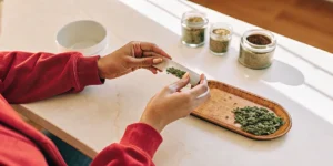 Hyper-realistic scene of a person's hands rolling a cannabis joint with ground material on a rolling paper, on a bright table with jars of cannabis.