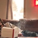 Tired man with dreadlocks lying on a brown couch with hand on face, a book and mug nearby.