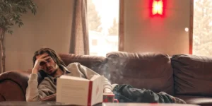 Tired man with dreadlocks lying on a brown couch with hand on face, a book and mug nearby.