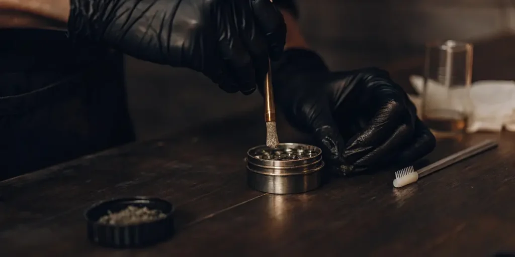 Gloved hands using a brush to clean a metal cannabis grinder on a dark wooden table.