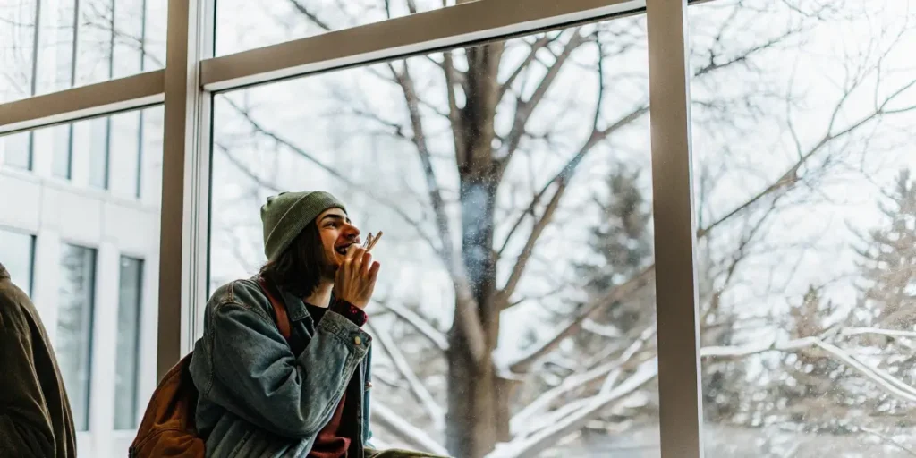 Young man smoking a joint while sitting by a window during a snowy winter day.