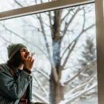 Young man smoking a joint while sitting by a window during a snowy winter day.