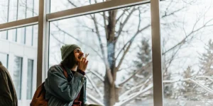 Young man smoking a joint while sitting by a window during a snowy winter day.
