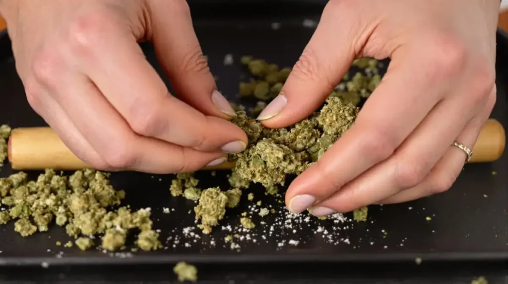 Hands breaking cannabis buds manually on a tray with a rolling pin in the background