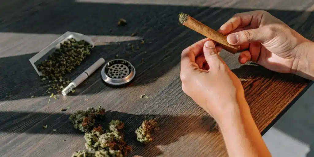 person rolling a cannabis blunt on a wooden table with grinder and buds.