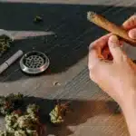 person rolling a cannabis blunt on a wooden table with grinder and buds.