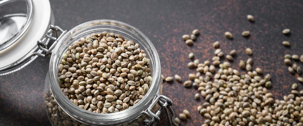 A glass jar filled with cannabis seeds, with some seeds scattered on a dark surface.