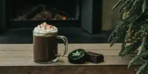 Steaming mug of cannabis-infused hot chocolate with whipped cream, next to a cannabis tin and brownie, by a fireplace and Christmas tree.
