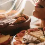Person holding a dark edible to their mouth, with a tray of assorted chocolates, dried fruits, and plums on a textured brown surface.