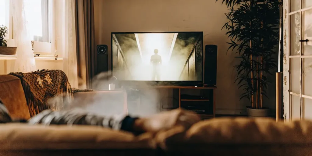 Person lying on a sofa with smoke, watching a TV showing a figure in a misty hallway.