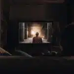 Person with back to camera watching a TV in a dark room, showing a figure in a tunnel.