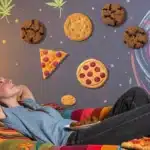 Woman reclining on a couch with floating cookies, pizza slices, and a psychedelic swirl backdrop featuring cannabis leaves.
