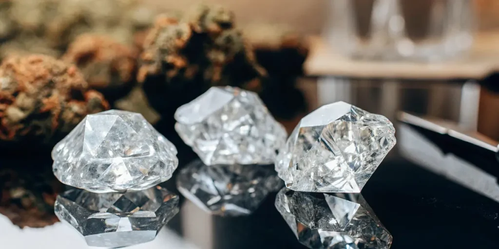 Two golden-hued THCA crystals reflecting on a light brown surface, with blurred cannabis in background.