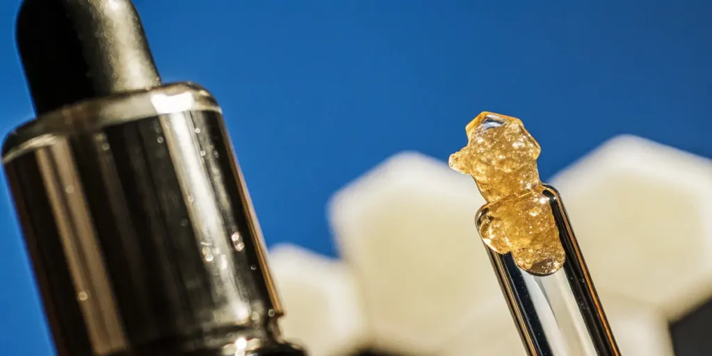 Hyper-realistic close-up of golden, crystalline THCA dabs on the tip of a glass dropper, with blurred white geometric shapes on a blue background.