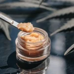 Hyper-realistic close-up of a metal dab tool scooping golden THCA badder from a glass jar, with blurred cannabis leaves in the background.