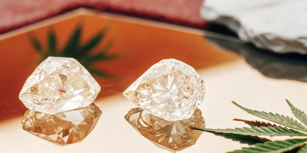 Two clear THCA crystals reflecting on a golden surface, beside a cannabis leaf.