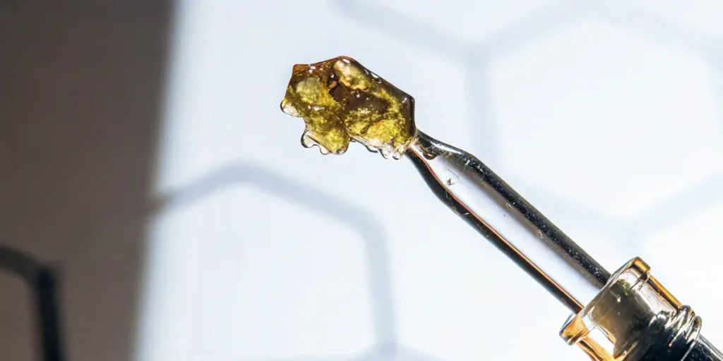 Hyper-realistic close-up of golden, crystalline THCA dabs on the tip of a glass dropper.