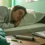 Person sleeping in bed with cannabis and vape pen on the nightstand, alongside a cup with visible smoke rising in a softly lit bedroom