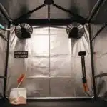 Gloved hand brushing a surface inside a grow tent, with "Hydrogen Peroxide," "Isopropyl Alcohol," and "Bleach" labels on the walls.