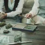 Two people rolling joints on a wooden table with cannabis buds, rolling papers, and grinders in a casual setting