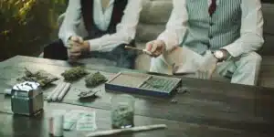 Two people rolling joints on a wooden table with cannabis buds, rolling papers, and grinders in a casual setting