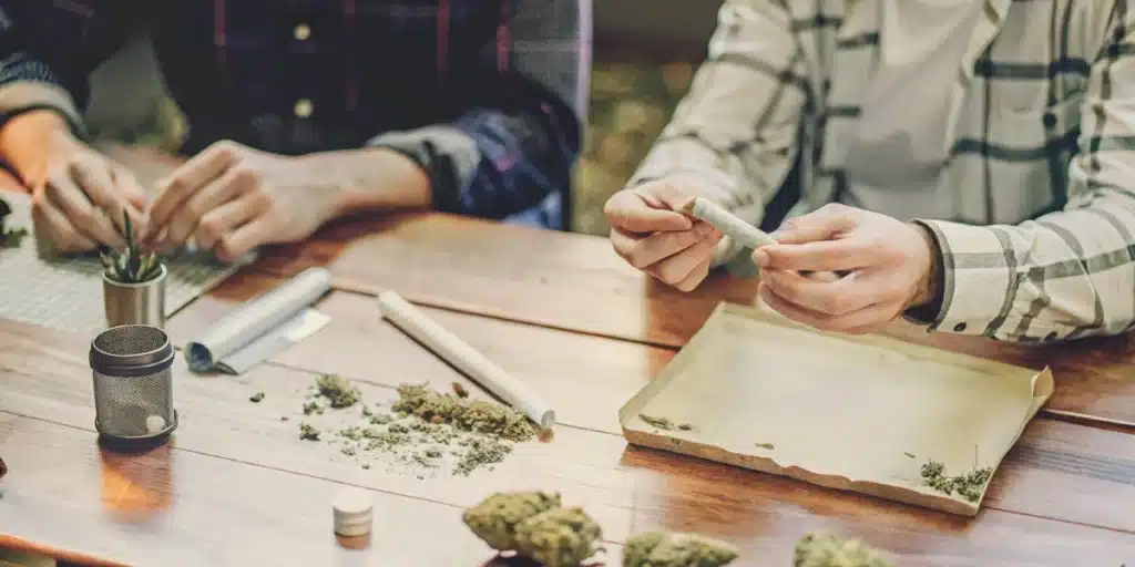 A person rolling a cannabis joint at a wooden table with scattered buds and rolling tools