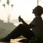 Silhouette of a person holding a cannabis oil bottle, relaxing on a mat on a sunny balcony with palm trees and water in the background.