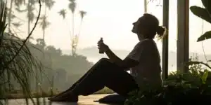 Silhouette of a person holding a cannabis oil bottle, relaxing on a mat on a sunny balcony with palm trees and water in the background.