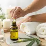Hand using a dropper to dispense CBD oil on a spa table, surrounded by rolled towels, lit candles, and cannabis leaves