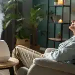 Person relaxing in a recliner with eyes closed, as an aroma diffuser emits mist on a side table with a dropper bottle, and a blurred plant and candles in the background.