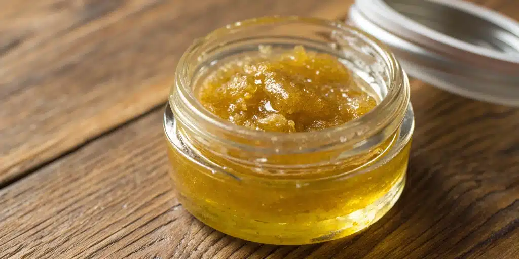 Close-up of CBD Sugar Wax in a jar with natural lighting and cannabis leaves on a wooden surface.
