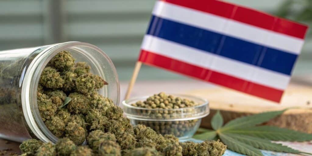 A glass jar spilling cannabis buds next to a Thai flag, cannabis seeds, and a hemp leaf.