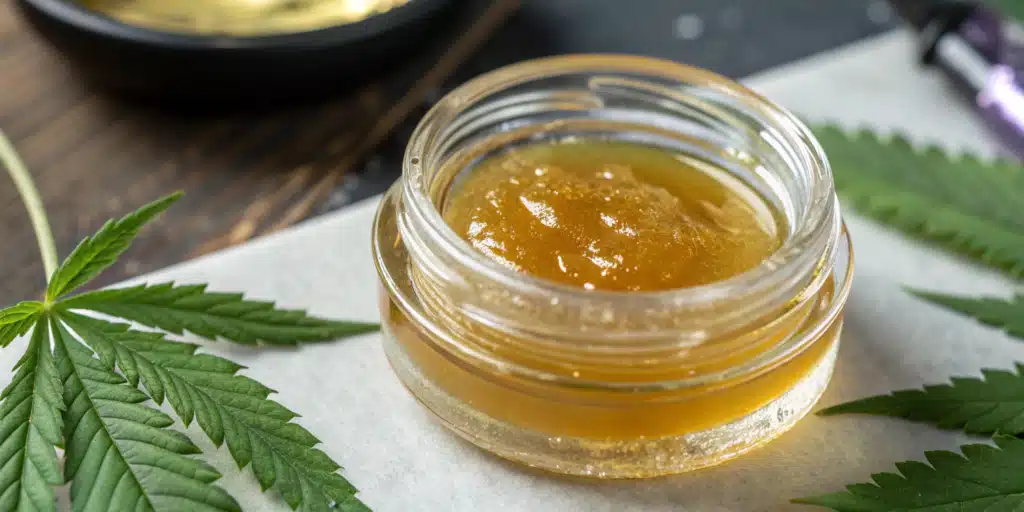Golden CBD concentrate in a glass jar surrounded by green cannabis leaves on a white surface.