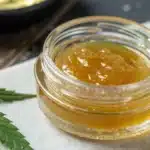 Golden CBD concentrate in a glass jar surrounded by green cannabis leaves on a white surface.