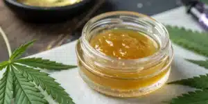 Golden CBD concentrate in a glass jar surrounded by green cannabis leaves on a white surface.