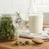 Glass jar of cannamilk recipe with hemp buds, measuring cup, and milk carton on wooden counter.
