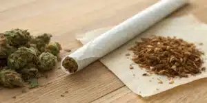 Cannabis joint next to loose tobacco and buds on a wooden surface.