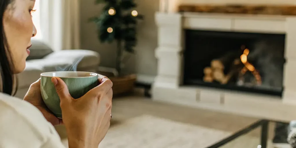 A woman sipping green tea from a cup while sitting by a cozy fireplace.