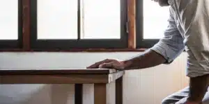 Close-up of a man's hand gripping a wooden table edge, next to a sunlit window.