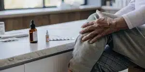 Close-up of an elderly person touching their aching knee with CBD drops and medication placed on a counter.