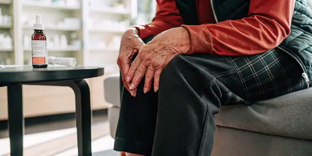 Elderly person holding their painful knee while a CBD tincture bottle sits on a nearby table in a clinical setting.