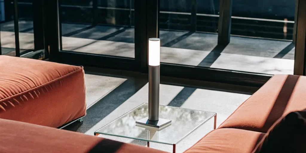 Modern minimalist table lamp glowing on a glass coffee table in a bright living room.
