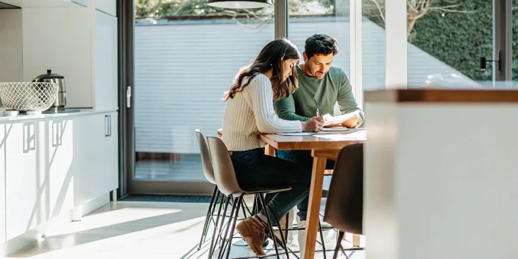A couple sitting at a wooden table in a bright modern kitchen reviewing paperwork together.