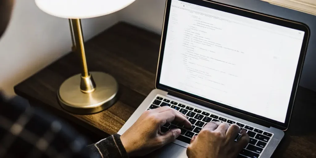 Close-up of hands typing programming code on a laptop beside a desk lamp.